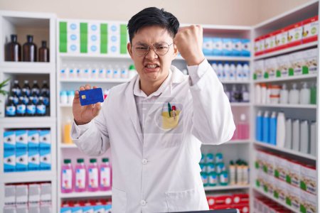 Foto de Young asian man working at pharmacy drugstore holding credit card annoyed and frustrated shouting with anger, yelling crazy with anger and hand raised - Imagen libre de derechos