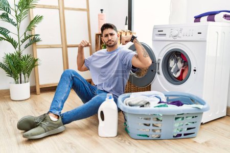 Téléchargez les photos : Young hispanic man putting dirty laundry into washing machine showing arms muscles smiling proud. fitness concept. - en image libre de droit