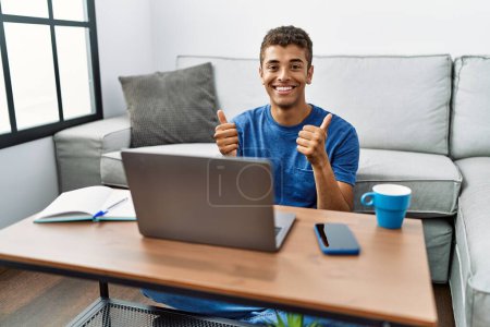 Photo for Young handsome hispanic man using laptop sitting on the floor success sign doing positive gesture with hand, thumbs up smiling and happy. cheerful expression and winner gesture. - Royalty Free Image