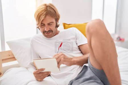 Foto de Joven hombre rubio escribiendo en el cuaderno acostado en la cama en el dormitorio - Imagen libre de derechos