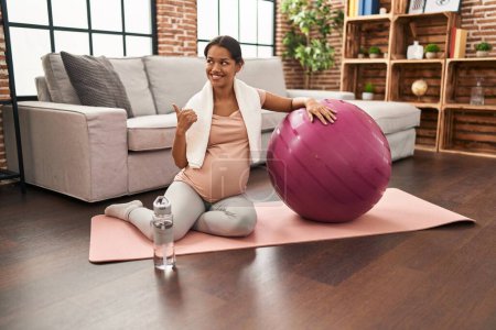 Foto de Joven embarazada sentada en esterilla de yoga con bola de pilates apuntando el pulgar hacia un lado sonriendo feliz con la boca abierta - Imagen libre de derechos