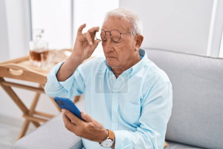 Foto de Hombre de pelo gris senior usando un teléfono inteligente sentado en el sofá en casa - Imagen libre de derechos