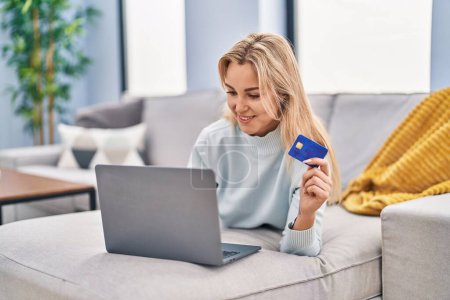 Foto de Mujer rubia joven usando el ordenador portátil y la tarjeta de crédito tumbado en el sofá en casa - Imagen libre de derechos