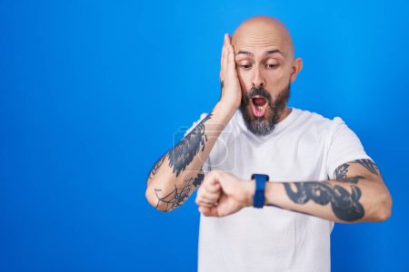 Téléchargez les photos : Homme hispanique avec des tatouages debout sur fond bleu regardant le temps de la montre inquiet, peur de se faire tard - en image libre de droit