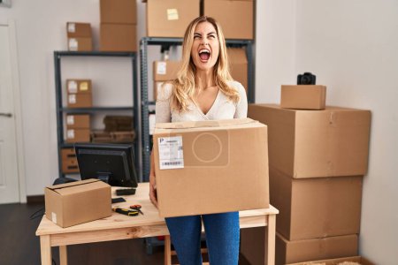 Photo for Young blonde woman working at small business ecommerce holding big box angry and mad screaming frustrated and furious, shouting with anger looking up. - Royalty Free Image
