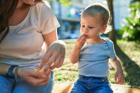 Photo for Mother and son sitting on bench together eating little worms snack at park - Royalty Free Image