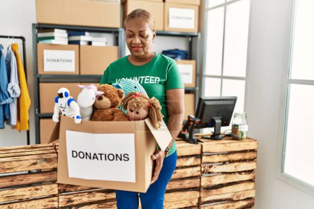 Photo for Senior african american woman wearing volunteer uniform holding toys donations package at charity center - Royalty Free Image