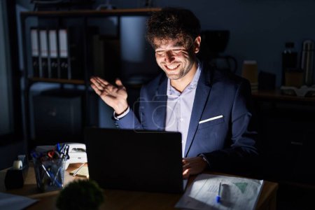 Photo for Hispanic young man working at the office at night smiling cheerful presenting and pointing with palm of hand looking at the camera. - Royalty Free Image
