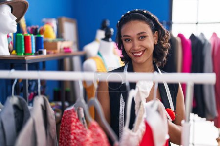 Téléchargez les photos : Jeune belle femme hispanique tailleur souriant confiant tenant des vêtements sur rack à l'usine de vêtements - en image libre de droit