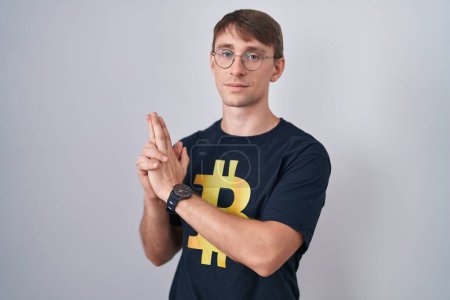 Téléchargez les photos : Caucasian blond man wearing bitcoin t shirt holding symbolic gun with hand gesture, playing killing shooting weapons, angry face - en image libre de droit
