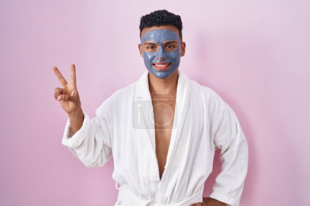 Téléchargez les photos : Jeune homme hispanique portant un masque de beauté et peignoir de bain souriant regardant la caméra montrant des doigts faisant signe de victoire. numéro deux. - en image libre de droit