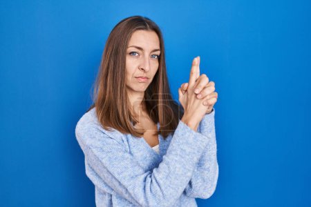 Foto de Mujer joven de pie sobre fondo azul sosteniendo un arma simbólica con gesto de mano, jugando a matar armas de fuego, cara enojada - Imagen libre de derechos