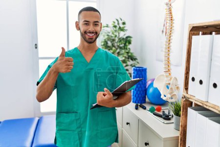 Photo for African american physiotherapist man working at pain recovery clinic doing happy thumbs up gesture with hand. approving expression looking at the camera showing success. - Royalty Free Image
