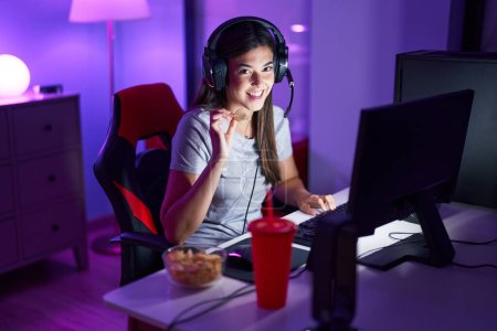 Foto de Joven hermosa mujer hispana serpenteante jugando videojuego comiendo patatas fritas en la sala de juegos - Imagen libre de derechos