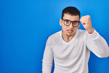 Téléchargez les photos : Young arab man wearing casual white shirt and glasses angry and mad raising fist frustrated and furious while shouting with anger. rage and aggressive concept. - en image libre de droit