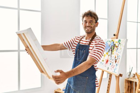 Téléchargez les photos : Jeune homme hispanique souriant confiant tenant toile au studio d'art - en image libre de droit