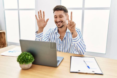 Foto de Joven hombre guapo con barba trabajando en la oficina usando computadora portátil mostrando y señalando con los dedos número siete mientras sonríe confiado y feliz. - Imagen libre de derechos