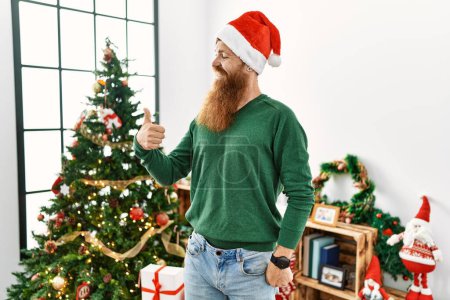 Téléchargez les photos : Homme rousse à longue barbe portant un chapeau de Noël par arbre de Noël regardant fier, souriant en faisant le pouce vers le haut geste sur le côté - en image libre de droit