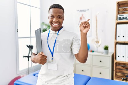 Téléchargez les photos : Jeune homme africain de physiothérapie travaillant à la clinique de récupération de la douleur souriant heureux pointant avec la main et le doigt vers le côté - en image libre de droit