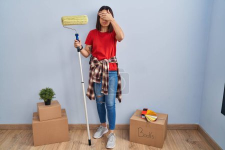 Foto de Mujer hispana joven pintando paredes de casa con rodillo de pintura cubriendo los ojos con la mano, luciendo seria y triste. concepto de ocultamiento y rechazo sin visión - Imagen libre de derechos
