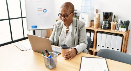 Foto de Trabajadora de negocios afroamericana mayor usando laptop y tomando café en la oficina - Imagen libre de derechos