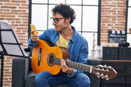 Foto de Joven músico hispano tocando guitarra clásica usando smartphone en estudio de música - Imagen libre de derechos