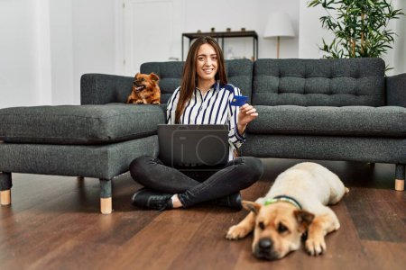 Téléchargez les photos : Jeune femme hispanique utilisant ordinateur portable et carte de crédit assis sur le sol avec des chiens à la maison - en image libre de droit