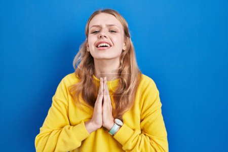 Téléchargez les photos : Young caucasian woman standing over blue background begging and praying with hands together with hope expression on face very emotional and worried. begging. - en image libre de droit