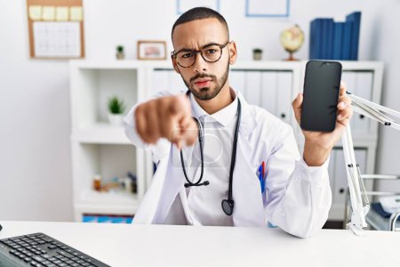 Photo for African american doctor man holding smartphone showing blank screen at the clinic pointing with finger to the camera and to you, confident gesture looking serious - Royalty Free Image