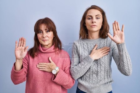 Foto de Mother and daughter standing over blue background swearing with hand on chest and open palm, making a loyalty promise oath - Imagen libre de derechos