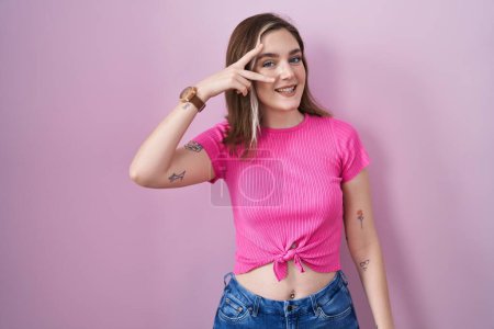 Photo for Blonde caucasian woman standing over pink background doing peace symbol with fingers over face, smiling cheerful showing victory - Royalty Free Image