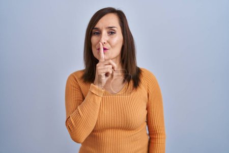 Foto de Middle age brunette woman standing wearing orange sweater asking to be quiet with finger on lips. silence and secret concept. - Imagen libre de derechos