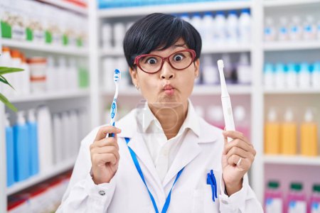 Téléchargez les photos : Young asian woman with short hair doing toothbrush comparative at pharmacy making fish face with mouth and squinting eyes, crazy and comical. - en image libre de droit