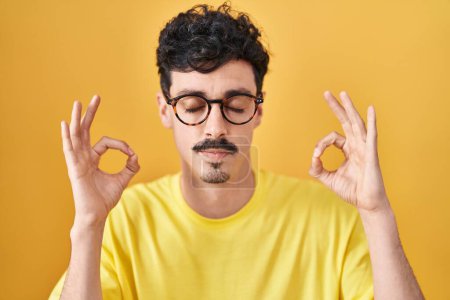Téléchargez les photos : Hispanic man wearing glasses standing over yellow background relax and smiling with eyes closed doing meditation gesture with fingers. yoga concept. - en image libre de droit