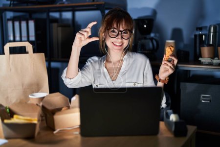 Photo for Young beautiful woman working using computer laptop and eating delivery food smiling and confident gesturing with hand doing small size sign with fingers looking and the camera. measure concept. - Royalty Free Image