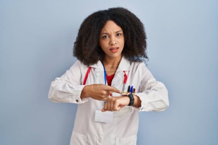 Foto de Joven mujer afroamericana vistiendo uniforme médico y estetoscopio con prisa apuntando a ver el tiempo, la impaciencia, molesto y enojado por retraso plazo - Imagen libre de derechos