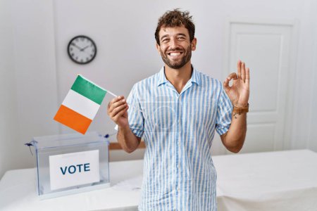 Foto de Joven hombre guapo en las elecciones de campaña política la celebración de bandera de Irlanda haciendo signo bien con los dedos, sonriendo gesto amistoso excelente símbolo - Imagen libre de derechos