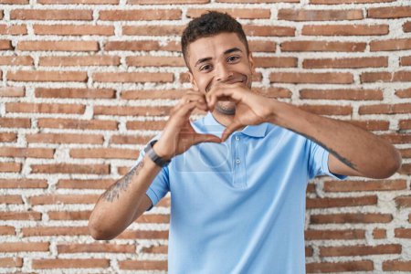 Téléchargez les photos : Jeune homme brésilien debout sur un mur de briques souriant dans l'amour faisant forme de symbole de coeur avec les mains. concept romantique. - en image libre de droit