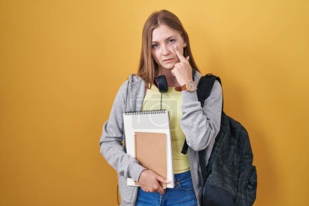 Photo for Young caucasian woman wearing student backpack and holding books pointing to the eye watching you gesture, suspicious expression - Royalty Free Image
