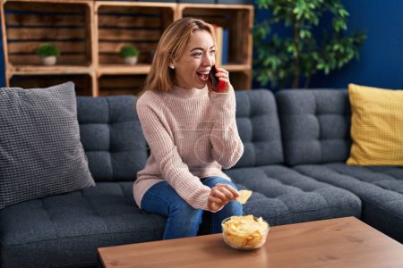 Photo for Young woman talking on smartphone eating chips potatoes at home - Royalty Free Image