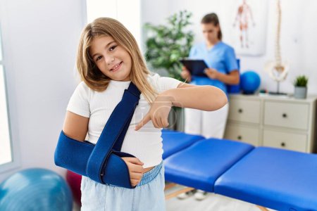 Photo for Blonde little girl wearing arm on sling at rehabilitation clinic smiling happy pointing with hand and finger - Royalty Free Image
