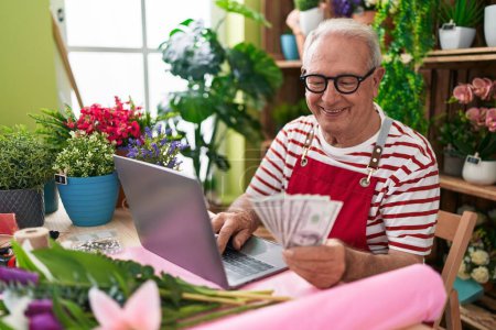 Foto de Floristería de hombre de pelo gris de mediana edad usando portátil contando dólares en floristería - Imagen libre de derechos