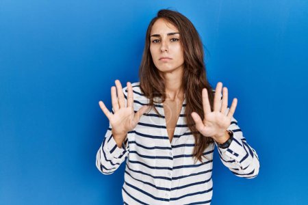 Foto de Mujer hispana joven de pie sobre fondo azul aislado alejando las palmas de las manos mostrando rechazo y negación con miedo y expresión repugnante. parada y prohibido. - Imagen libre de derechos