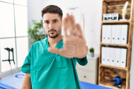 Photo for Young physiotherapist man working at pain recovery clinic doing stop sing with palm of the hand. warning expression with negative and serious gesture on the face. - Royalty Free Image