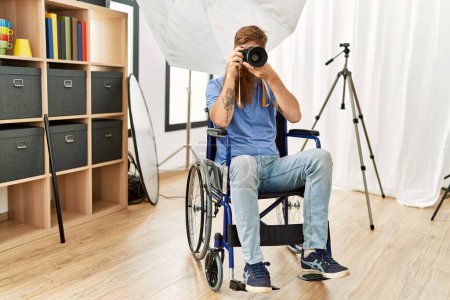 Foto de Joven fotógrafo pelirrojo sentado en silla de ruedas usando cámara profesional en el estudio de fotografía - Imagen libre de derechos