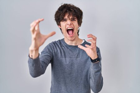 Foto de Young man standing over isolated background shouting frustrated with rage, hands trying to strangle, yelling mad - Imagen libre de derechos