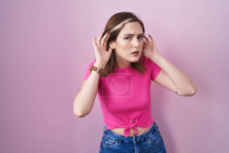 Foto de Mujer rubia caucásica de pie sobre fondo rosa tratando de escuchar ambas manos en el gesto de la oreja, curioso por chismes. problema auditivo, sordo - Imagen libre de derechos