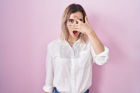 Photo for Young beautiful woman standing over pink background peeking in shock covering face and eyes with hand, looking through fingers with embarrassed expression. - Royalty Free Image