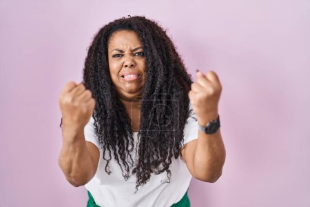 Téléchargez les photos : Plus size hispanic woman standing over pink background angry and mad raising fists frustrated and furious while shouting with anger. rage and aggressive concept. - en image libre de droit