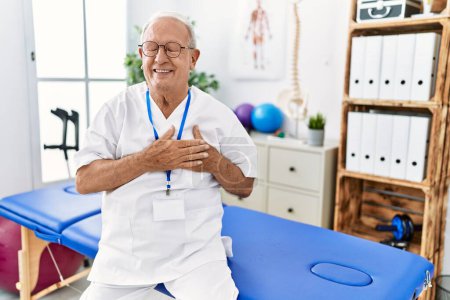 Photo for Senior physiotherapy man working at pain recovery clinic smiling with hands on chest with closed eyes and grateful gesture on face. health concept. - Royalty Free Image
